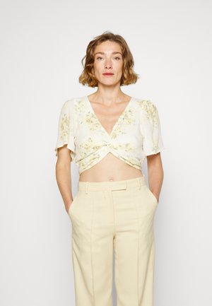 Top corto branco com estampado floral e mangas curtas e volumosas, apresentando uma frente torcida. Combinado com calças beiges de cintura alta, enfatizando um caimento descontraído.