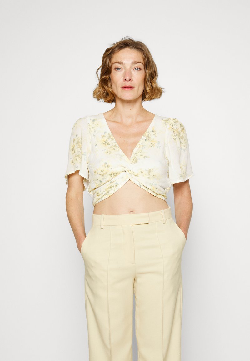 Top corto branco com estampado floral e mangas curtas e volumosas, apresentando uma frente torcida. Combinado com calças beiges de cintura alta, enfatizando um caimento descontraído.
