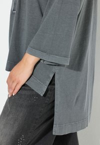 Graues, weit geschnittenes Oberteil mit Dreiviertelärmeln und seitlichen Schlitzen. Weicher Stoff mit leicht strukturiertem Finish. Kombiniert mit dunkelblauen Jeans mit minimalen Akzenten.