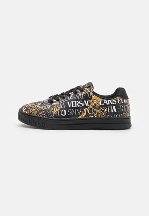 Sort lavtop sneakers med gyldent barokmønster, hvid "VERSACE JEANS COUTURE" tekst, sorte snørebånd og præget branding på sålen.