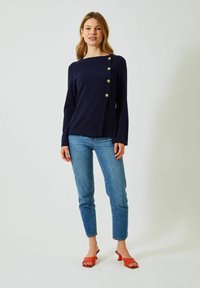 Marinblå stickad topp med båturringning, långa ärmar, asymmetrisk design och gula knappar längs ena axeln, kombinerad med ljusblå jeans och röda skor.
