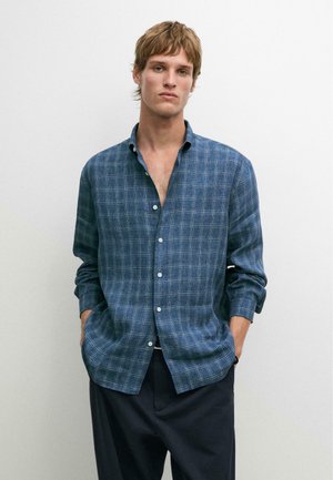 Jeune homme portant une chemise à carreaux bleue déboutonnée et un pantalon sombre, debout les mains dans les poches devant un fond blanc uni.