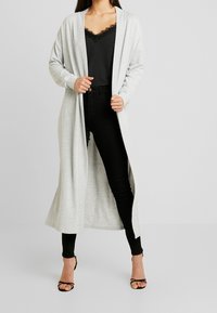 Lichtgrijze lange cardigan gemaakt van zachte stof, met een open voorkant en lange mouwen, gecombineerd met een zwart topje en aansluitende broek.
