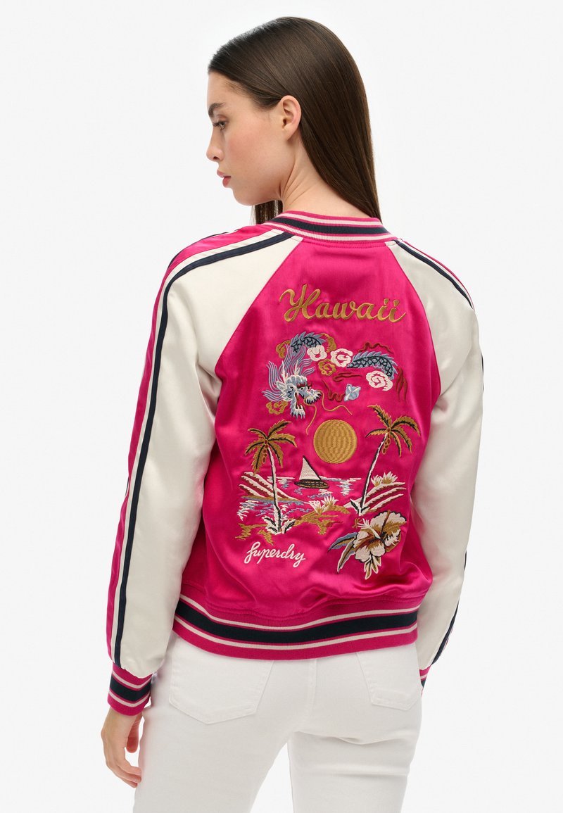 Veste bomber satinée, rose avec des manches blanches. Présente un motif brodé représentant Hawaï, des palmiers, un soleil et un bateau. Poignets et ourlet côtelés.