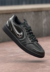 Crne i sive tenisice s mrežastim i kožnim detaljima, kromirani Nike swoosh, crne vezice, suspendirane iznad betonske površine.