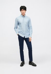 Camisa azul clara com botões e bolso no peito, combinada com calças azul-marinho e sapatos pretos. Textura suave, corte ajustado e design clássico.