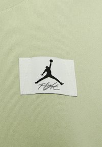 Tejido acanalado verde claro con una etiqueta blanca que presenta un logo Jumpman negro y el texto "Flight", cosida en la prenda.