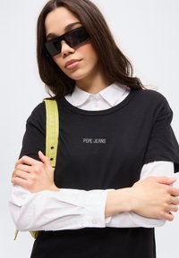 Νεαρή γυναίκα που φοράει μαύρα γυαλιά ηλίου, λευκό πουκάμισο με κουμπιά κάτω από μαύρο t-shirt Pepe Jeans, κρατώντας έναν κίτρινο υφασμάτινο ιμάντα στον ώμο.