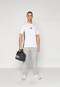 Λευκό κοντομάνικο μπλουζάκι με μαύρο λογότυπο adidas και πλαϊνές ρίγες, γκρι φόρμα, και μαύρο σάκο duffle με λευκό λογότυπο.