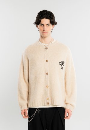 Jeune homme portant un cardigan beige boutonné avec un logo noir brodé sur la poitrine et un pantalon noir avec un accessoire chaîne.