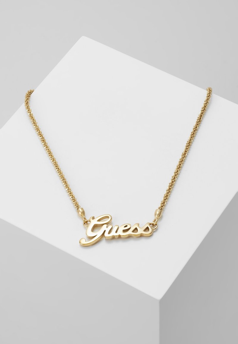 Collier chaîne en or avec un pendentif inscrit « Guess » en écriture cursive sur une surface blanche inclinée.