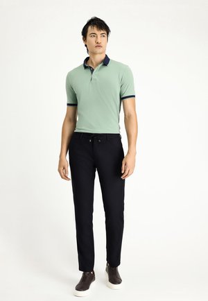 Hellgrünes Poloshirt mit marineblauem Kragen und Besatz, kombiniert mit schwarzen Hosen und dunkelbraunen Slippern. Glatte Textur, figurnahe Passform.