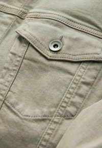 Camisa de algodón beige claro con un bolsillo con solapa, que presenta un botón de metal en tono plateado, costuras visibles y un detalle de tela texturizada.