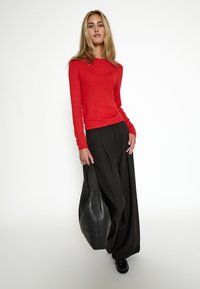 Top de manga larga rojo acanalado combinado con pantalones negros holgados. La modelo sostiene un gran bolso de cuero negro, con textura suave y forma redondeada.