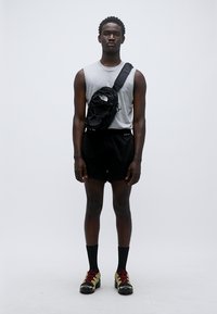 Μαύρη τσάντα χιαστί, γκρι αμάνικο πουκάμισο, μαύρες βερμούδες και αθλητικά παπούτσια. Λείο ύφασμα, μινιμαλιστικός σχεδιασμός και ορατό λογότυπο της μάρκας στην τσάντα.