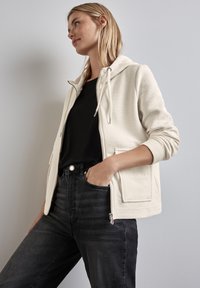 Felpa zip con cappuccio in crema chiaro, realizzata con un tessuto testurizzato, cappuccio regolabile e tasche frontali, abbinata a jeans neri a vita alta.