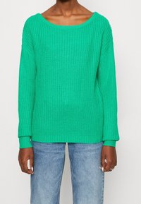 Grön stickad tröja med bred båtkrage, ribbade ärmar och nederkant, samt en lös passform, i kombination med ljusblå denimjeans.