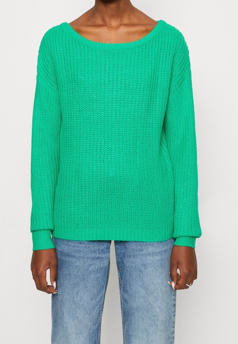 Grön stickad tröja med bred båtkrage, ribbade ärmar och nederkant, samt en lös passform, i kombination med ljusblå denimjeans.