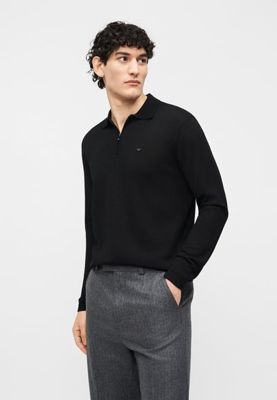 Άνδρας που φοράει μαύρο μακρυμάνικο πόλο με φερμουάρ, μπαίνει μέσα σε γκρι παντελόνι, στέκεται με το ένα χέρι στην τσέπη, κοιτάζοντας αριστερά.