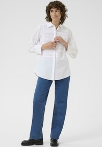 Chemise blanche à boutons avec panneau avant texturé, manches longues et ourlet arrondi, associée à un jean bleu coupe droite et des chaussures noires.