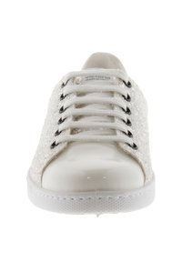 Vit låg sneaker med glittriga sidor, vita snören trätt genom svarta ögla, och glänsande tåspets, sedd framifrån.