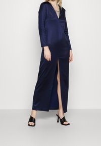 Navy satin long dress with long sleeves and a deep V-neck, featuring a thigh-high slit. Worn with black open-toe heels.