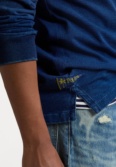 Mörkblå långärmad skjorta med ribbade ärmslut, sidoflikar och en gul logotypetikett med en palm vid nederkanten.