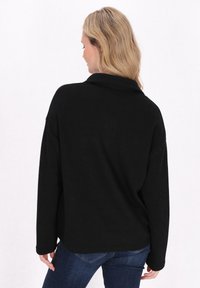Fekete, oversize, hosszú ujjú felső, texturált anyagból, galléros nyakkal és laza szabással, kék farmernadrággal viselve.