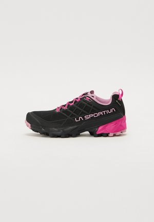 Zapatilla de running negra con acentos en rosa. Presenta un upper texturizado, suela acolchada, un diseño de suela agresivo y una lengüeta en el talón.