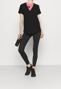 T-shirt noir enroulé par-dessus une chemise rose, associé à un legging noir. Le mannequin tient une bouteille d'eau noire et porte des chaussures de sport noires.