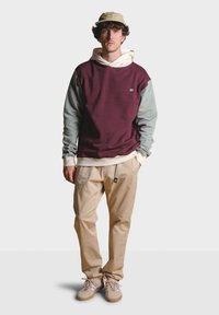 Bordový a šedý sveter s bielou mikinou pod ním, skombinovaný s béžovými nohavicami a svetlohnedými topánkami. Doplnky zahŕňajú svetlú čiapku.