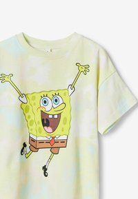 T-shirt jaune tie-dye avec un grand personnage de dessin animé de forme carrée. Manches courtes, col rond et un design ludique avec deux couleurs.