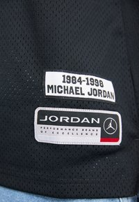 Zwarte netstof met kleine perforaties bevat twee gestikte labels: de ene geeft "1984-1998 MICHAEL JORDAN" aan, en de andere "JORDAN PERFORMANCE BRAND."