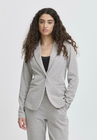 Šedý blazer s vzorem herringbone a s klopovým límečkem, dvěma předními kapsami a jedním knoflíkem, v kombinaci s odpovídajícími kalhotami.