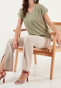 Top a V di colore verde oliva con texture, abbinato a pantaloni beige a zampa d'elefante con fessure laterali. Calzature includono tacchi a fascia multicolore. Sedia in legno.