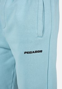 Šviesiai mėlynos sportinės kelnės, pagamintos iš minkštos medžiagos, su siuvinėtu juodu "PEGADOR" logotipu šone ir šoninėmis kišenėmis.