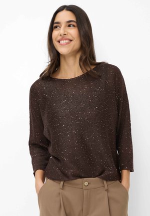 Femme souriante aux longs cheveux bruns portant un pull en maille brun foncé moucheté et un pantalon taille haute beige, les mains dans les poches.