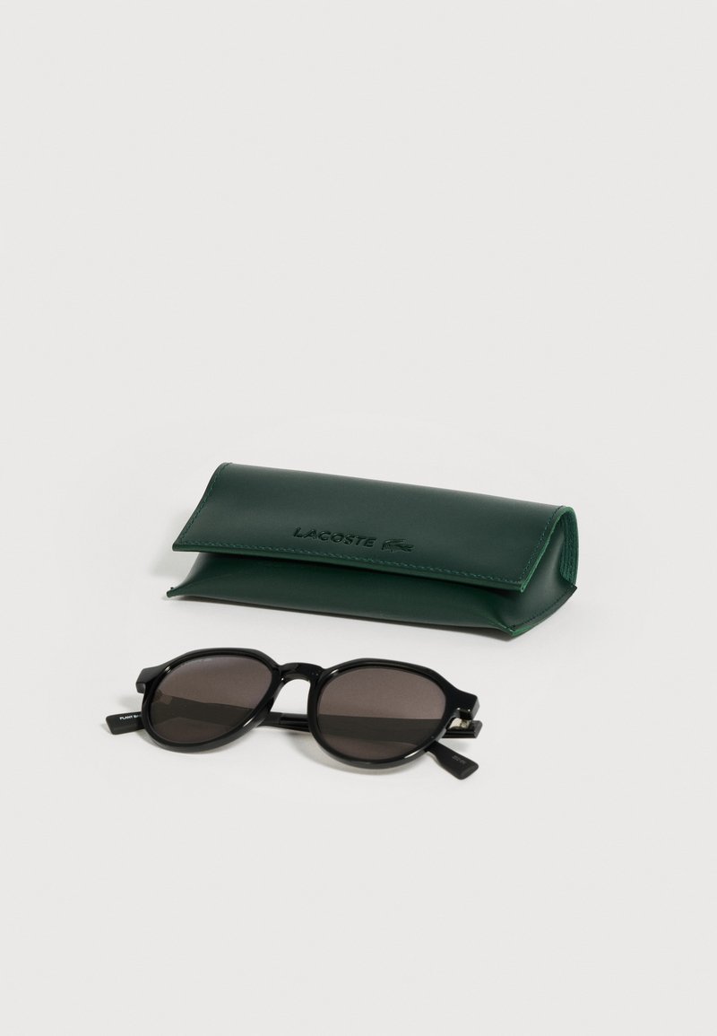 Black oval sunglasses with dark lenses placed next to a closed dark green Lacoste branded eyeglass case on a white background.