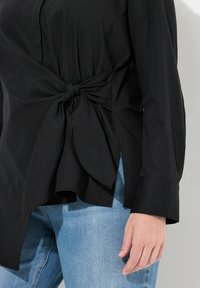 Camisa negra con detalle de cintura anudada, mangas largas y aberturas laterales; confeccionada en tejido suave, combinada con unos vaqueros azul claro.