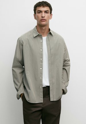 Uomo con camicia larga a righe abbottonata sopra una t-shirt bianca e pantaloni scuri, in piedi contro uno sfondo chiaro e uniforme con le mani in tasca.