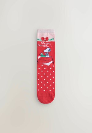 Punainen sukka, jossa on valkoisia pilkkuja, vaaleanpunainen kantti ja kuvio, jossa on koira ja lahja tekstillä "Rakas Joulupukki". Materiaali vaikuttaa pehmeältä ja joustavalta.