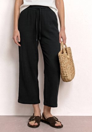 Femme portant un pantalon large texturé noir et des sandales marron à boucle, tenant un sac à main en paille tressée contre un mur blanc.