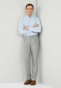 Camicia azzurro chiaro con bottoni, pantaloni grigi a plissettatura, e scarpe in suede marroni. In piedi contro uno sfondo neutro, con le braccia incrociate.