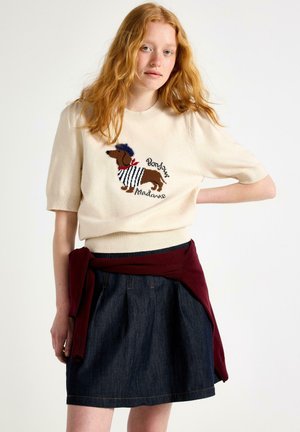 Kvinde med langt rødt hår iført en cremet sweater med et hundemotiv og en marineblå nederdel, med en mørkerød sweater bundet om livet.