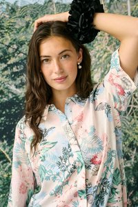 Jeune femme aux longs cheveux ondulés portant une chemise à imprimé floral, des boucles d'oreilles en perles et un chouchou noir, posant avec la main sur la tête devant un arrière-plan feuillu.