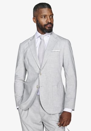 Uomo con barba che indossa un completo grigio chiaro, camicia bianca e cravatta abbinata, guarda verso sinistra con una mano in tasca.