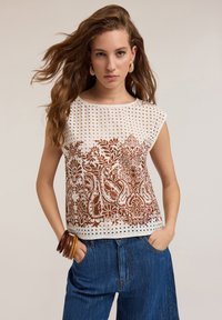 Top bianco a maniche corte con motivo traforato, caratterizzato da stampa floreale e paisley marrone. Abbinato a pantaloni di jeans blu a vita alta e bracciali di legno.