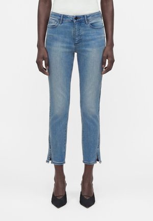 Eestvaade inimesest, kes kannab helerohelisi pahklukani teksapükse külgpoolsete lõhikutega ja musti teravate kontsadega kingi, seistes ühevärvilise tausta ees.