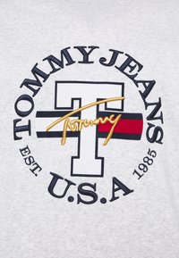 Šedé bavlněné tričko s velkým vyšívaným logem: "TOMMY JEANS," stylizované "T" a "USA" s červenými a námořnickými pruhy, založeno v roce 1985.