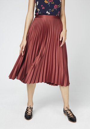Femme portant une jupe midi plissée couleur rouille, un haut sans manches à motif floral foncé, et des chaussures plates noires à lacets.